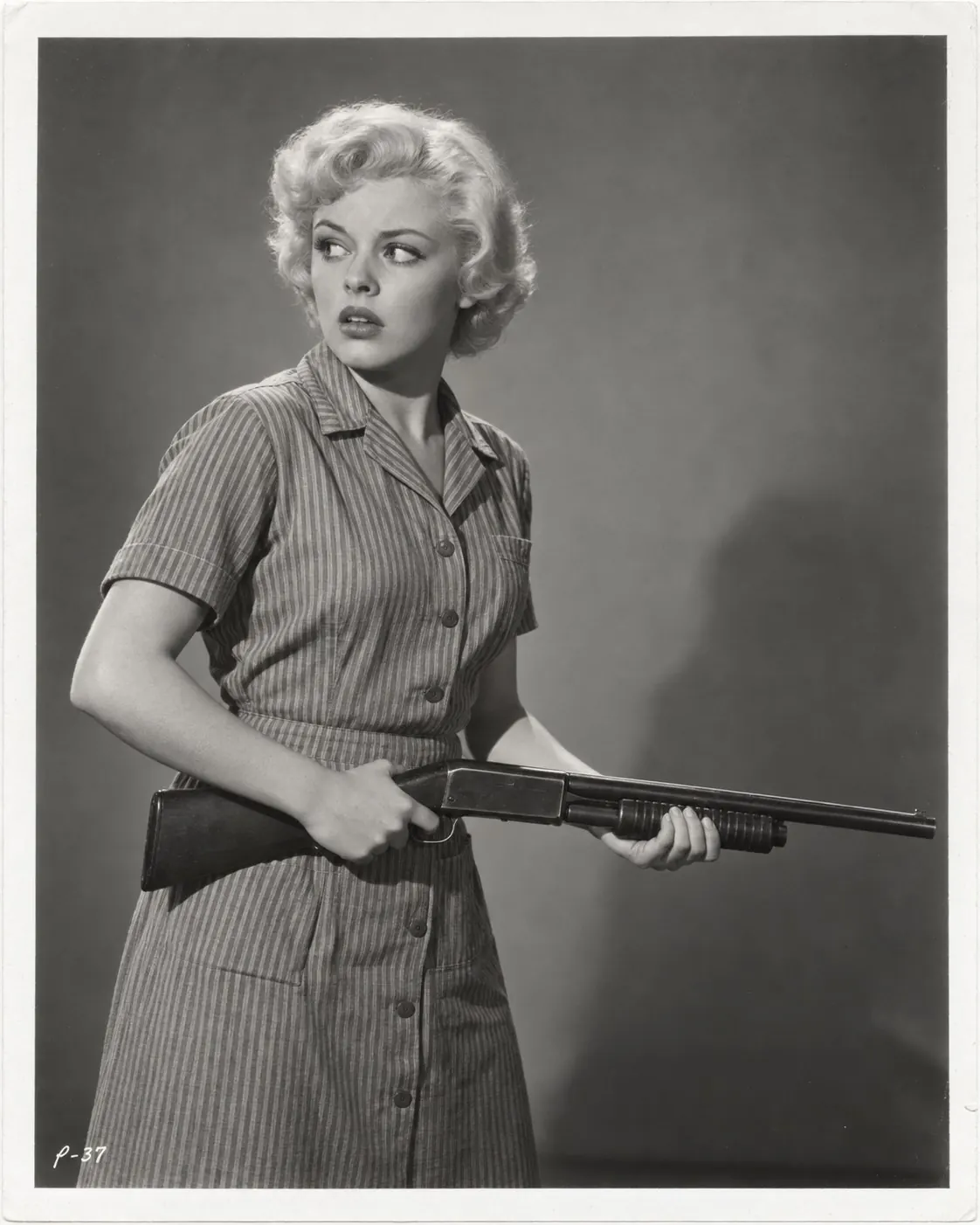 A vintage-style studio still of a woman in a striped dress holding a shotgun