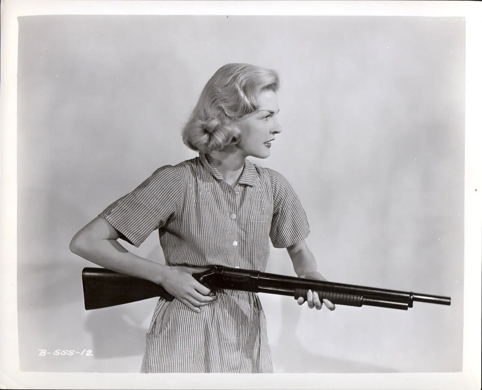 A real 1955 studio still of an actress in a striped dress holding a shotgun