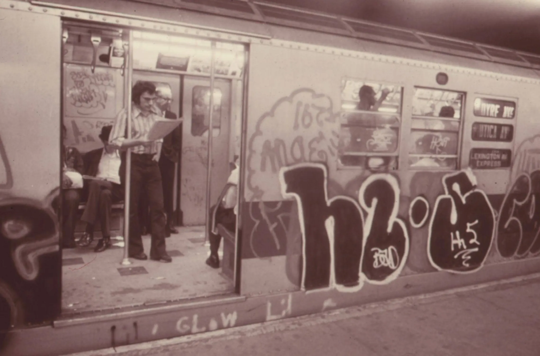 A graffiti-covered Lexington Avenue express car with its doors open, a man reading a newspaper inside, 1974